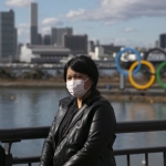 Seorang turis yang mengenakan topeng berpose untuk foto dengan cincin Olimpiade di latar belakang, di distrik Odaiba Tokyo. foto: theguardian