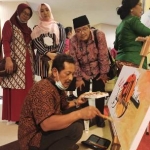 Drs. Romadlon Sukardi (jongkok pakai kopiah hitam) menyaksikan salah satu seniman yang sedang mendemonstrasikan kebolehannya dalam melukis. foto: ist