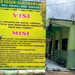 Kepala SDN Barengkrajan 1, Drs. Sunandar M.Pd. berdiri di depan visi misi sekolah.